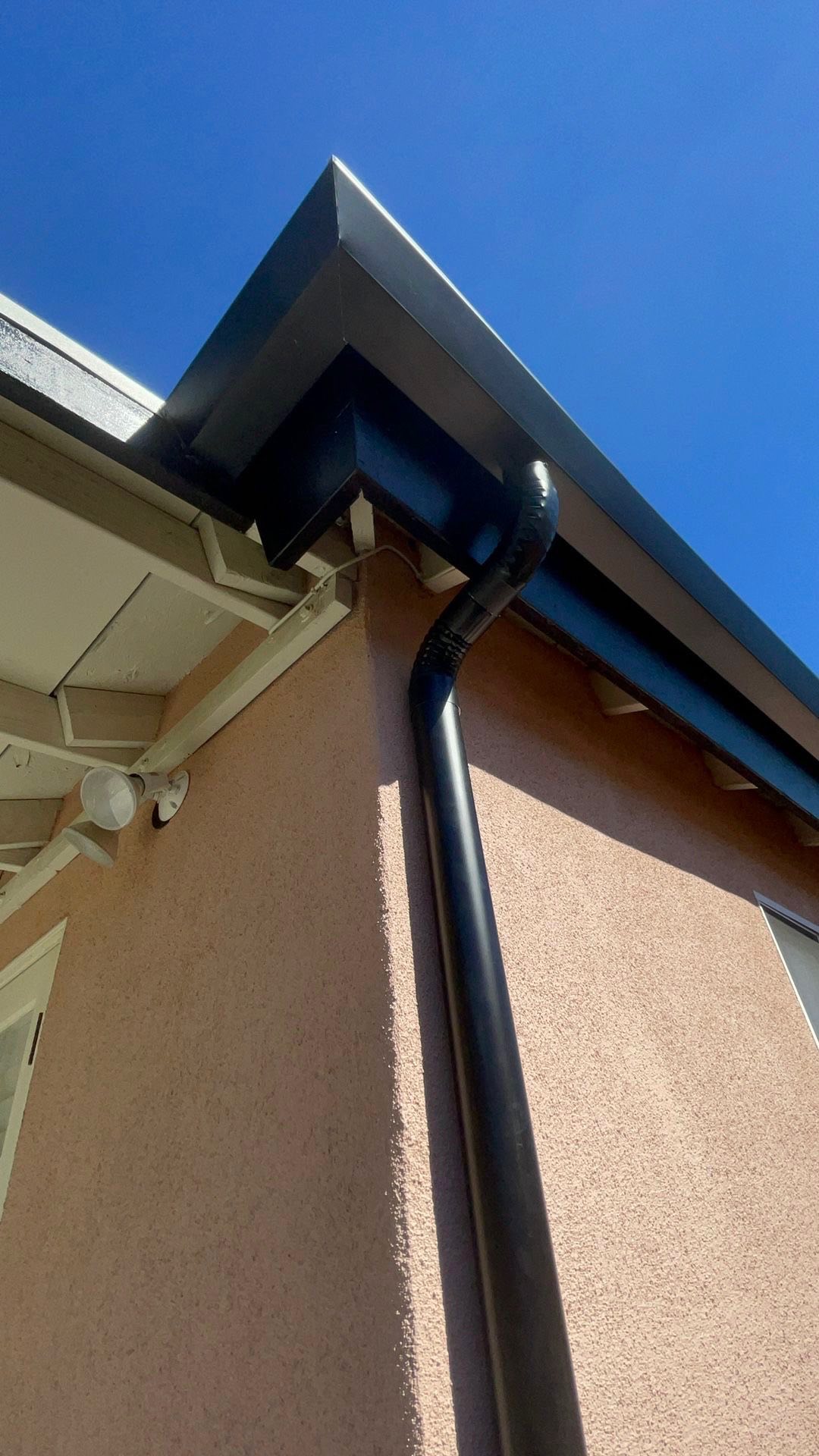 Black corrugated round downspout below a dark gutter on a stucco home in the Los Angeles area